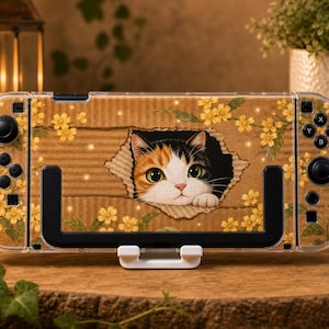 May include: A handheld gaming console featuring a protective skin with a brown cardboard texture background, yellow flowers, and a calico cat peeking through a torn hole in the center. The device has black buttons and joysticks on both sides.