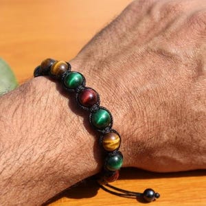 May include: A beaded bracelet featuring alternating round tiger eye and green cat eye stones, secured with black cord in a macrame style knotting pattern. The bracelet includes a sliding closure with two small black beads at the ends.