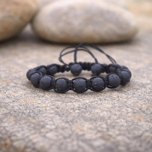 Peut inclure: Un bracelet en pierre de lave noire avec un cordon noir réglable. Le bracelet est composé de perles rondes et poreuses enfilées ensemble. L'arrière-plan comprend de grosses pierres lisses et une surface texturée.