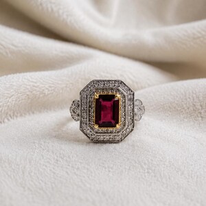 May include: A ring featuring a rectangular deep red gemstone set in a yellow gold bezel, surrounded by a double halo of small white stones set in a silver-toned metal frame with decorative side details on the band.
