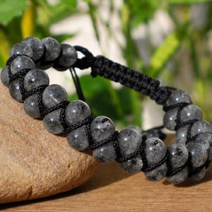 Pode incluir: Uma pulseira com duas filas paralelas de contas redondas cinza-escuras salpicadas, unidas por um cordão preto trançado. O design inclui um fecho de macramé preto ajustável com pontas de cordão pendentes.