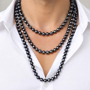 May include: A multi-strand necklace featuring three layers of smooth, dark gray, spherical beads. The beads have a reflective, metallic luster and are strung together on a dark cord, resting against a crisp, white collared button-down shirt.