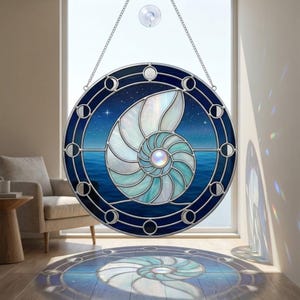 Könnte beinhalten: Rundes Buntglasfenster mit einem Nautilus-Muschel-Design. Das Kunstwerk hat ein blau-weißes Farbschema mit einem Sternenhimmel und einer Meereslandschaft. Das Fenster ist mit einem dunkelblauen Rahmen und Mondphasen-Details versehen.