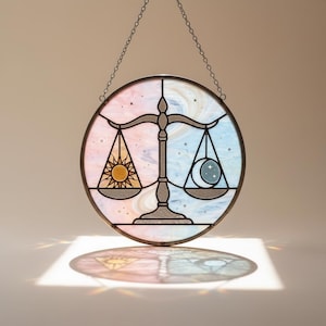 Può includere: Suncatcher rotondo in vetro colorato con un design a bilancia della Bilancia. Il design è diviso in due metà, una con un sole e l'altra con una luna e stelle. Lo sfondo è un mix di rosa, blu e bianco, con una catena per appenderlo.