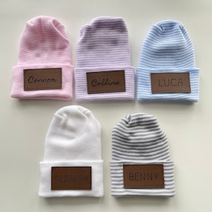 May include: Five striped baby hats with personalised leather name tags. The hats are in pink, purple, blue, white and grey.