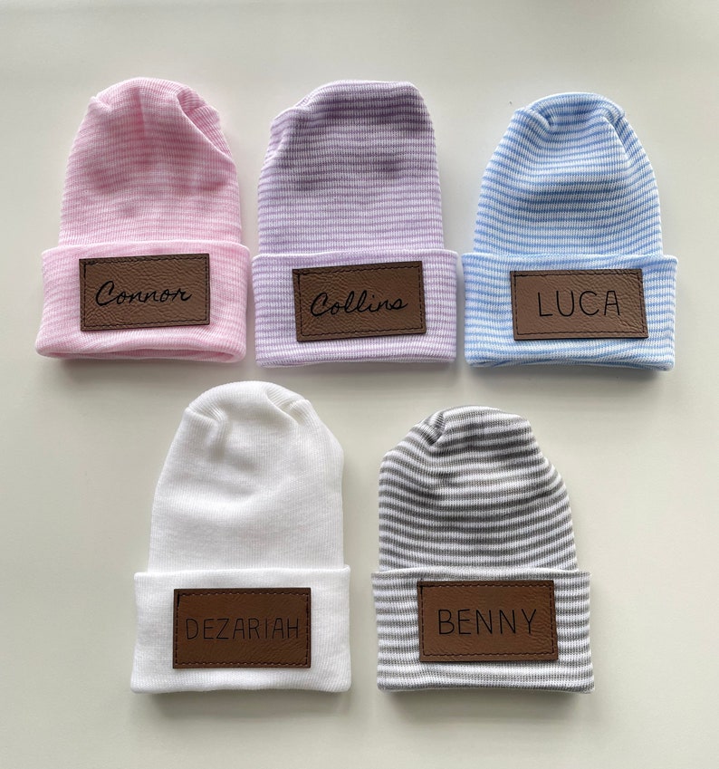 May include: Five striped baby hats with personalised leather name tags. The hats are in pink, purple, blue, white and grey.