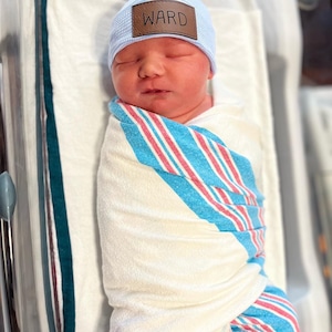 May include: A newborn baby wearing a light blue knitted hat with a brown leather patch that says "WARD" is swaddled in a white, blue and pink striped blanket.