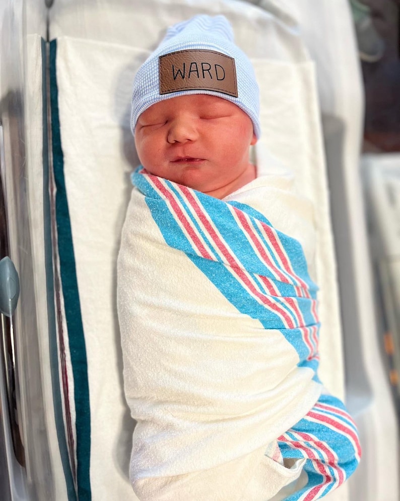 May include: A newborn baby wearing a light blue knitted hat with a brown leather patch that says "WARD" is swaddled in a white, blue and pink striped blanket.