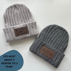May include: Two gray knitted beanies with brown leather patches. The patches have the names "Theo" and "Austin" written in black.