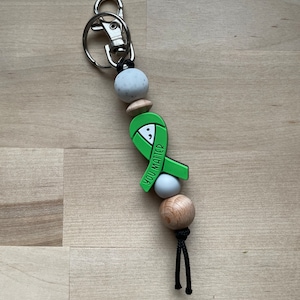 May include: Green ribbon keychain with the words "You Matter" on it. The keychain has a silver clasp and is made with wooden and speckled white beads.