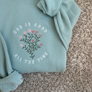 May include: A light blue sweatshirt with the text "God is good all the time" embroidered in white. The embroidery includes a floral design with pink flowers.
