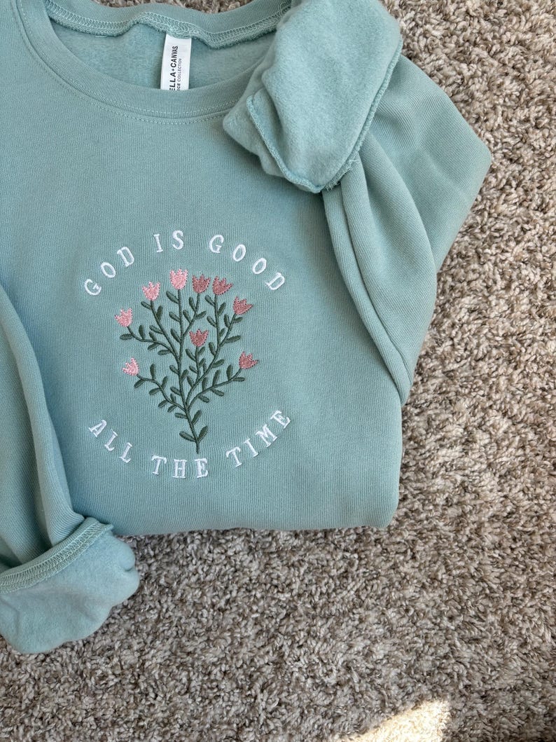 May include: A light blue sweatshirt with the text "God is good all the time" embroidered in white. The embroidery includes a floral design with pink flowers.