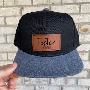 May include: Black and gray baseball cap with a brown leather patch that says "Foster MICHAEL" in black script.