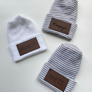 May include: Three white and grey striped knit hats with brown leather patches. The patches have the names "Kaspyan", "Beckett", and "Rhett" written on them.