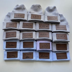 May include: A stack of 18 grey and white striped knitted hats with brown leather name tags. The names on the tags include: Levi, Leo, Baylor, Ari, Laken, Crew, Brooks, Lucas, Judah, Avery, Luca, Caden, Palushaj, Leo, Boden, Cooper Bradley, Roman Lee, Lucas, and Maverick.