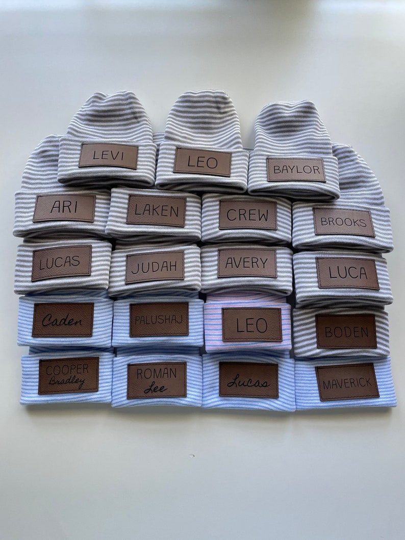 May include: A stack of 18 grey and white striped knitted hats with brown leather name tags. The names on the tags include: Levi, Leo, Baylor, Ari, Laken, Crew, Brooks, Lucas, Judah, Avery, Luca, Caden, Palushaj, Leo, Boden, Cooper Bradley, Roman Lee, Lucas, and Maverick.
