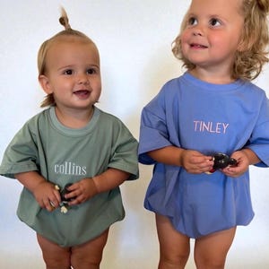 May include: A sage green short-sleeve t-shirt with the name collins printed in white and a periwinkle blue short-sleeve t-shirt with the name TINLEY printed in light pink. Both shirts feature a crew neckline and a loose, boxy fit design.
