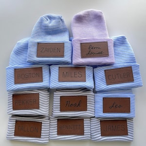 May include: Ten striped baby hats with personalised leather name tags. The hats are in shades of blue and grey. The names on the tags are Zaiden, Remi Louise, Boston, Miles, Cutler, Rekker, Noah, Leo, Fallon, Bennett, and James.