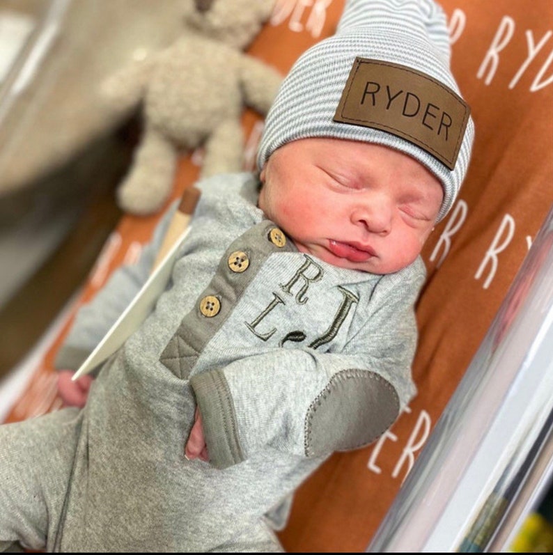 May include: A newborn baby wearing a grey and brown onesie with the letters "R.L." embroidered on the front. The baby is also wearing a grey and white striped beanie with a brown leather patch that says "RYDER".
