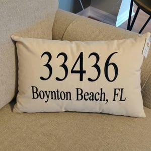 May include: Cream-colored rectangular pillow with the embroidered text "33436 Boynton Beach, FL" in navy blue. The pillow is placed on a beige upholstered armchair. A tag is attached to the pillow.