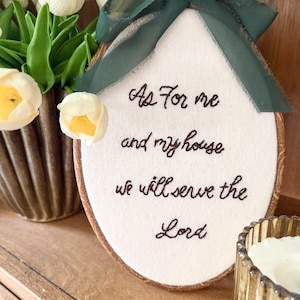 May include: An oval embroidery hoop containing white fabric with the dark brown embroidered text "As For me and my house we will serve the Lord." A dark green sheer ribbon is tied in a bow at the top of the wooden hoop frame.