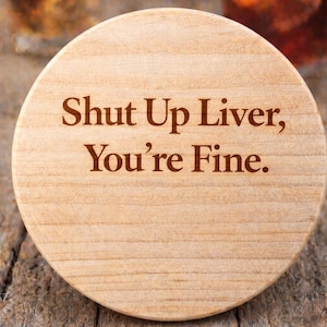 Puede incluir: Un posavasos redondo de madera clara con la frase "Shut Up Liver, You're Fine" grabada en marrón. El posavasos está grabado con láser y tiene el logotipo de Etsy.