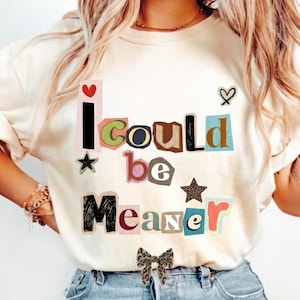 Puede incluir: Camiseta color crema con el texto "I Could Be Meaner" en estilo collage. El texto incluye un corazón rojo, una estrella negra y un lazo con estampado de leopardo. La camiseta se combina con unos vaqueros azules.