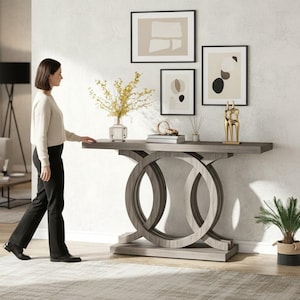 May include: A gray wooden console table with a unique double-circle base design. The table is adorned with decorative items, including framed art, a gold sculpture, and a vase of yellow flowers. A woman is standing next to the table.