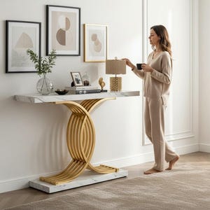 May include: A modern console table with a white marble top and a gold-colored, intertwined base. The table is placed against a white wall with framed artwork. A woman in a beige outfit stands nearby.