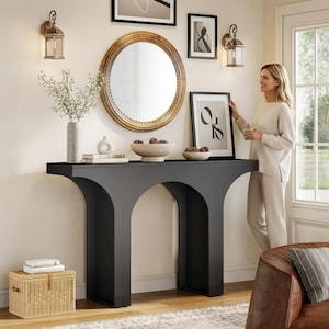 May include: A modern living room scene featuring a black console table with an arched base, a round gold-framed mirror, and framed artwork. Decorative elements include a vase with flowers, bowls, and sconces. A wicker basket and a brown leather chair are also visible.