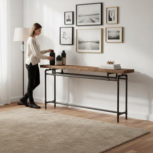 May include: A long, rustic wooden console table with black metal pipe legs. The table is adorned with black and beige vases, books, and decorative items. A gallery wall of framed black and white art hangs above the table. A woman is standing near the table.