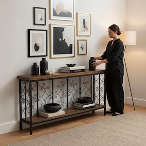 May include: A wooden console table with a black metal frame and decorative scrollwork. The table displays black vases, bowls, and books. Artwork in gold and black frames hangs on the white wall above the table.
