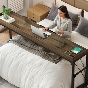 May include: A dark brown, rectangular bed desk with a black metal frame. The desk has a built-in tray and is positioned over a bed with white bedding. A laptop, tablet, and small plant are on the desk.