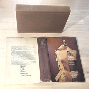 May include: An open book titled "Design Your Own Dress Patterns" by Adele P. Margolis, with a brown cover and a dress form on the right page. The book is on a light-colored surface, with the closed book visible above.