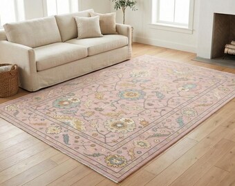 May include: A rectangular area rug featuring a soft pink background with a repeating floral pattern in muted tones of blue, cream, and tan. The rug has a decorative border and short fringe along the edges, displaying a classic, symmetrical design.