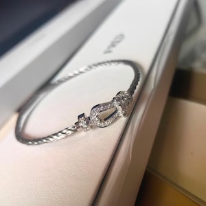 May include: A silver bracelet with a braided design. The bracelet features a horseshoe-shaped clasp adorned with small, sparkling stones. The bracelet is presented in a white box, suggesting a jewelry item.