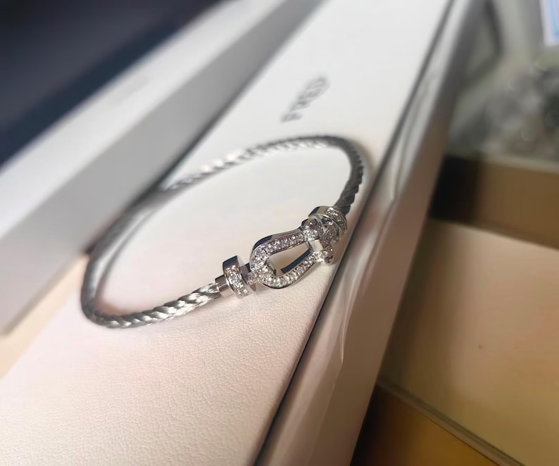 May include: A silver bracelet with a braided design. The bracelet features a horseshoe-shaped clasp adorned with small, sparkling stones. The bracelet is presented in a white box, suggesting a jewelry item.