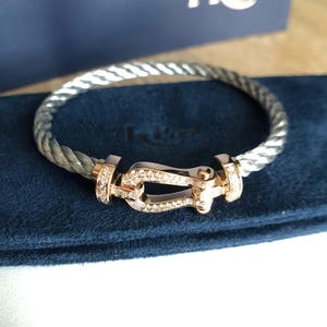 May include: A silver and grey twisted cable bracelet with a gold-coloured clasp. The clasp is encrusted with small, clear gemstones. The bracelet is resting on a dark blue surface.