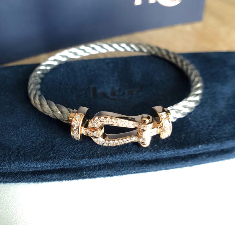 May include: A silver and grey twisted cable bracelet with a gold-coloured clasp. The clasp is encrusted with small, clear gemstones. The bracelet is resting on a dark blue surface.