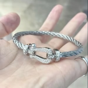 Könnte beinhalten: Ein silberfarbenes Armband mit geflochtenem Kabeldesign. Der Verschluss ist U-förmig und mit kleinen, klaren Edelsteinen besetzt. Das Armband wird in einer Hand gehalten, um die Details und das Design des Schmucks hervorzuheben.