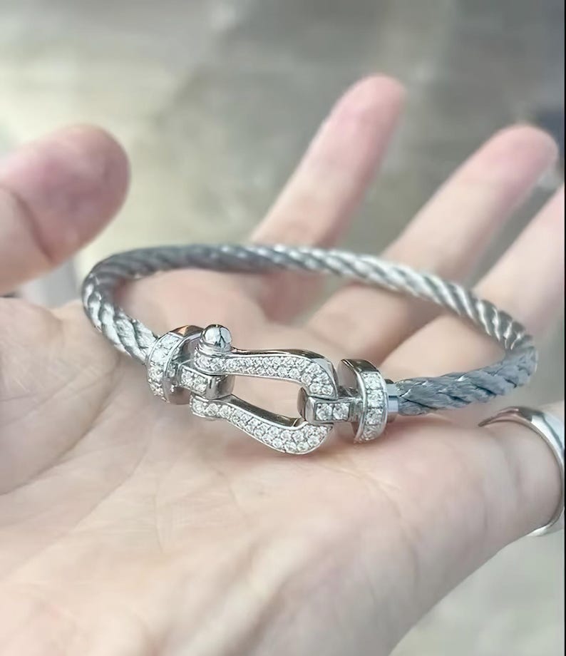 May include: A silver-toned bracelet with a braided cable design. The clasp is a U-shaped design, encrusted with small, clear gemstones. The bracelet is held in a hand, showcasing the jewelry's details and design.