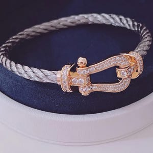 May include: A silver-coloured braided bracelet with a gold-coloured clasp. The clasp is decorated with small, clear gemstones. The bracelet is presented in a white and navy blue jewellery box.