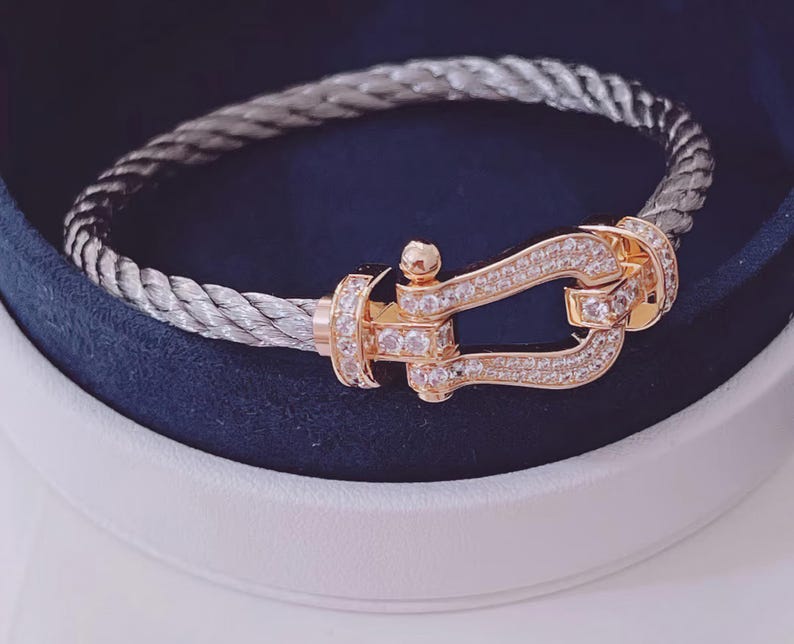 May include: A silver-coloured braided bracelet with a gold-coloured clasp. The clasp is decorated with small, clear gemstones. The bracelet is presented in a white and navy blue jewellery box.