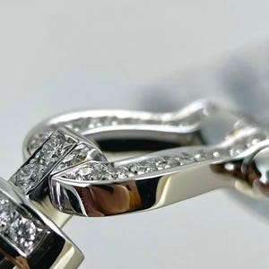 May include: Close-up of a silver-toned bracelet clasp with sparkling, clear gemstones. The clasp features a curved, rectangular design with a polished finish, and is set against a soft, blurred background.
