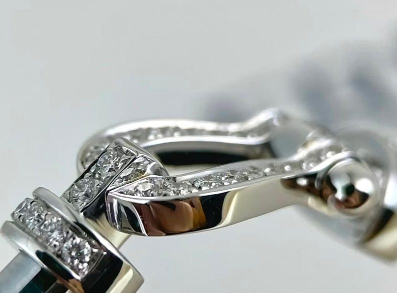 May include: Close-up of a silver-toned bracelet clasp with sparkling, clear gemstones. The clasp features a curved, rectangular design with a polished finish, and is set against a soft, blurred background.