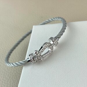 May include: A silver-toned bracelet with a cable design. The bracelet features a decorative clasp adorned with small, clear gemstones. The clasp is a focal point, adding sparkle to the jewelry. The bracelet is displayed on a white surface.