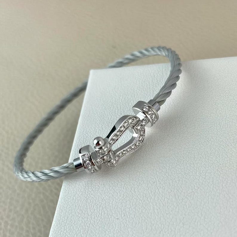 May include: A silver-toned bracelet with a cable design. The bracelet features a decorative clasp adorned with small, clear gemstones. The clasp is a focal point, adding sparkle to the jewelry. The bracelet is displayed on a white surface.