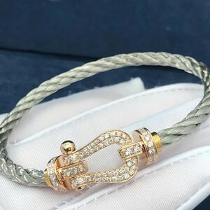 May include: A silver-toned braided bracelet with a rose gold clasp. The clasp is encrusted with small diamonds. The bracelet is displayed on a white surface, with a dark blue background.