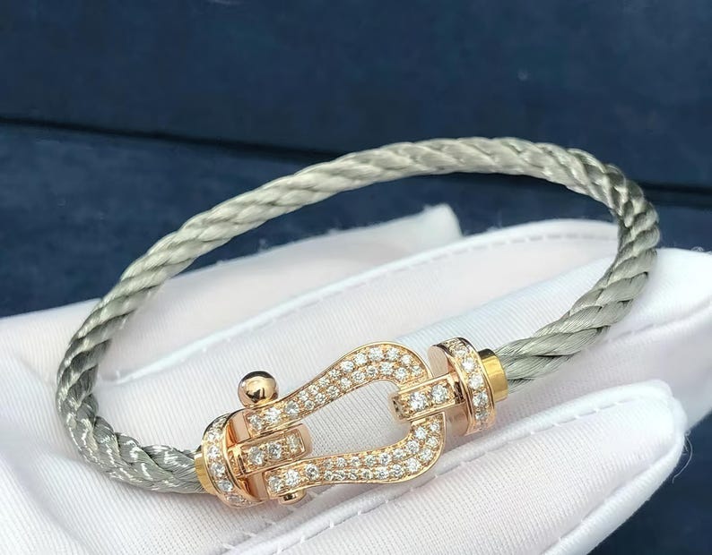 May include: A silver-toned braided bracelet with a rose gold clasp. The clasp is encrusted with small diamonds. The bracelet is displayed on a white surface, with a dark blue background.