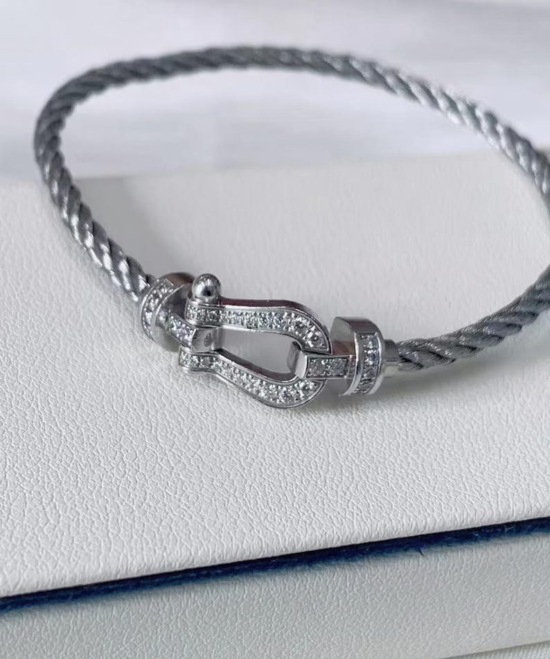 May include: A silver-colored bracelet with a braided cable design. The bracelet features a decorative clasp adorned with small, sparkling gemstones. The clasp is in the shape of a horseshoe. The bracelet is displayed on a white surface.